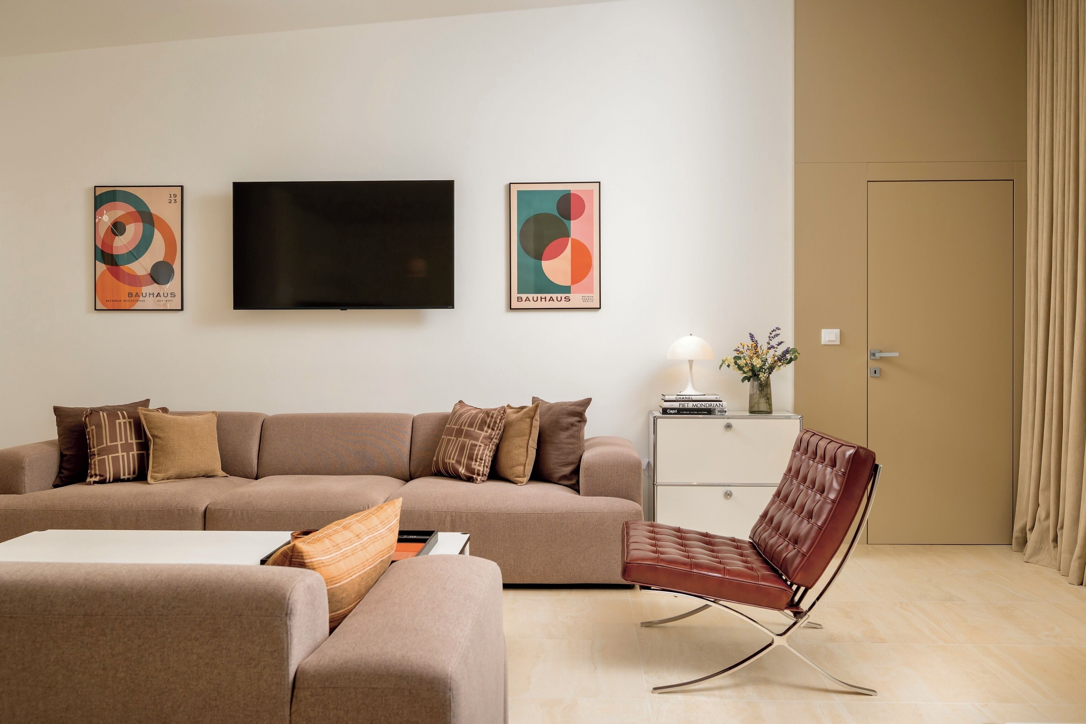 Uma sala de estar moderna com um sofá, uma poltrona de couro marrom, uma TV de tela plana e duas obras de arte na parede inspiradas no estilo Bauhaus.