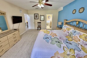 Une chambre à coucher comprenant un lit, une commode, un téléviseur, un ventilateur de plafond et une porte donnant sur une autre pièce.
