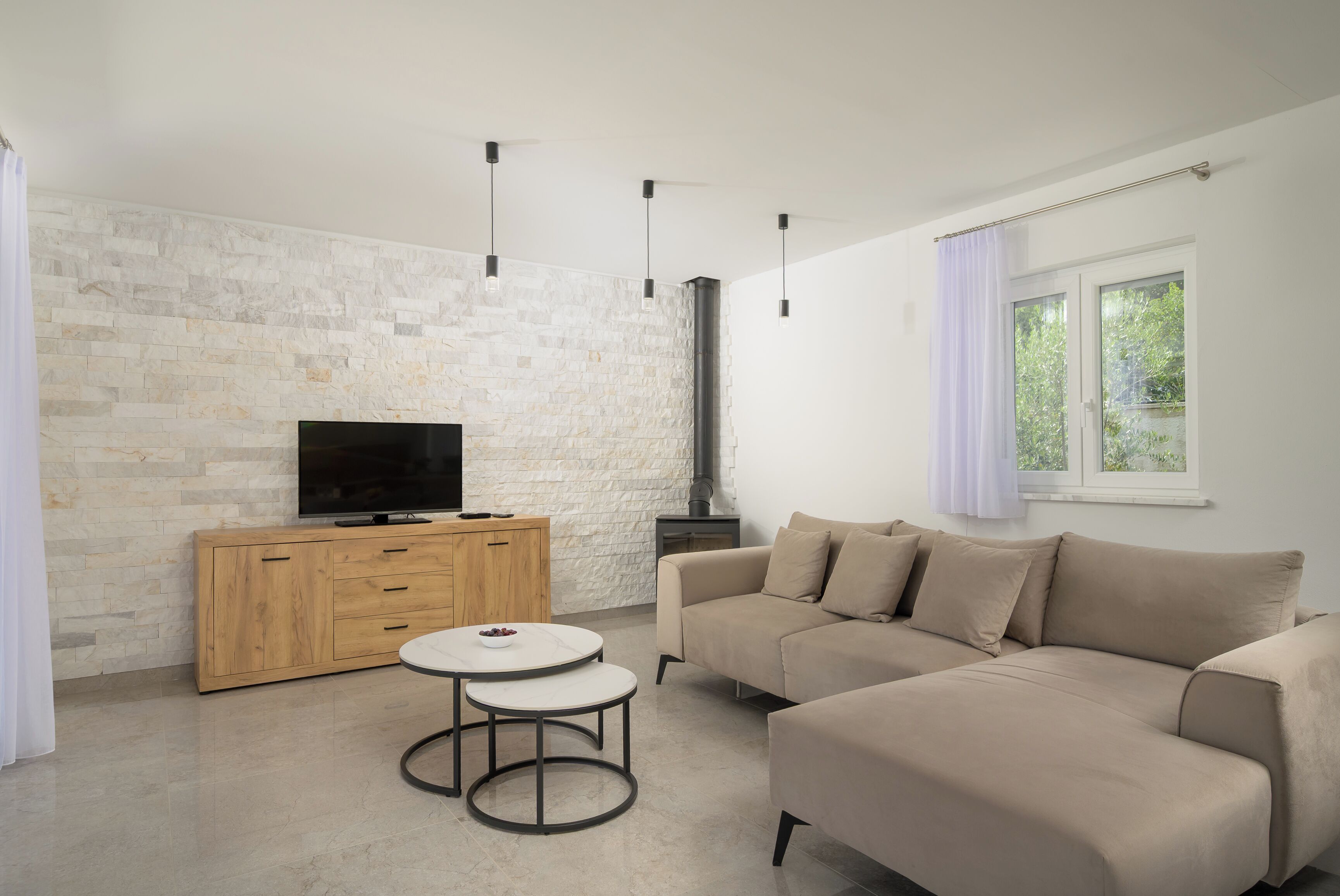 Un salon moderne avec un mur en pierre, un téléviseur à écran plat, un meuble en bois, un canapé beige et une table ronde.