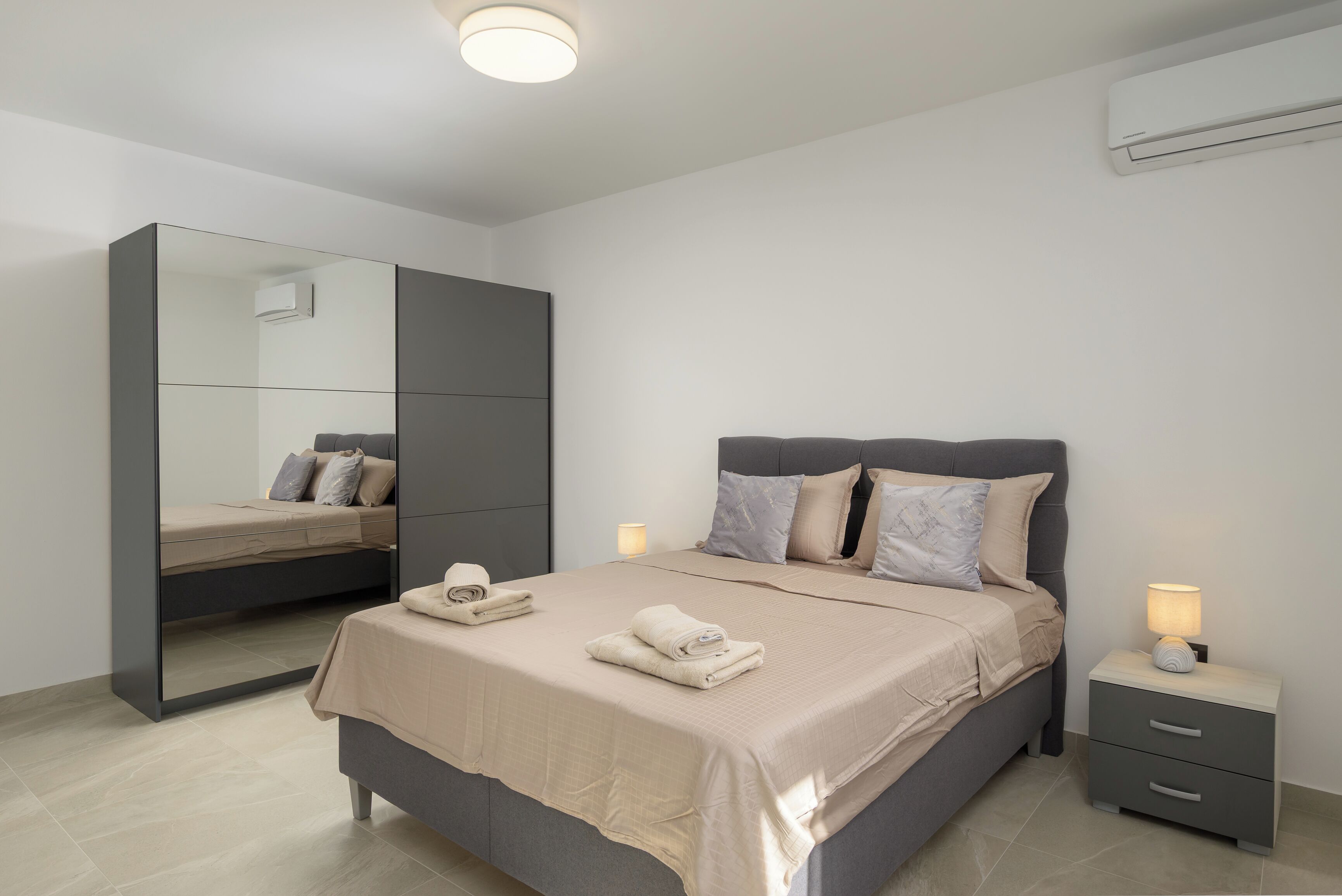 A modern bedroom with a bed, bedside tables, a mirror, and an air conditioning unit.