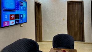 Ein Konferenzraum mit einem Flachbildfernseher, der verschiedene Apps zeigt, zwei dunklen Stühlen, einem Holztisch mit Rosenblüten und einer Decke mit Einbaustrahlern.