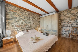 Ein Schlafzimmer mit Steinwand, Holzbalken, einem Bett mit weißer Decke, einem Nachttisch mit Lampe und einem Kleiderschrank.