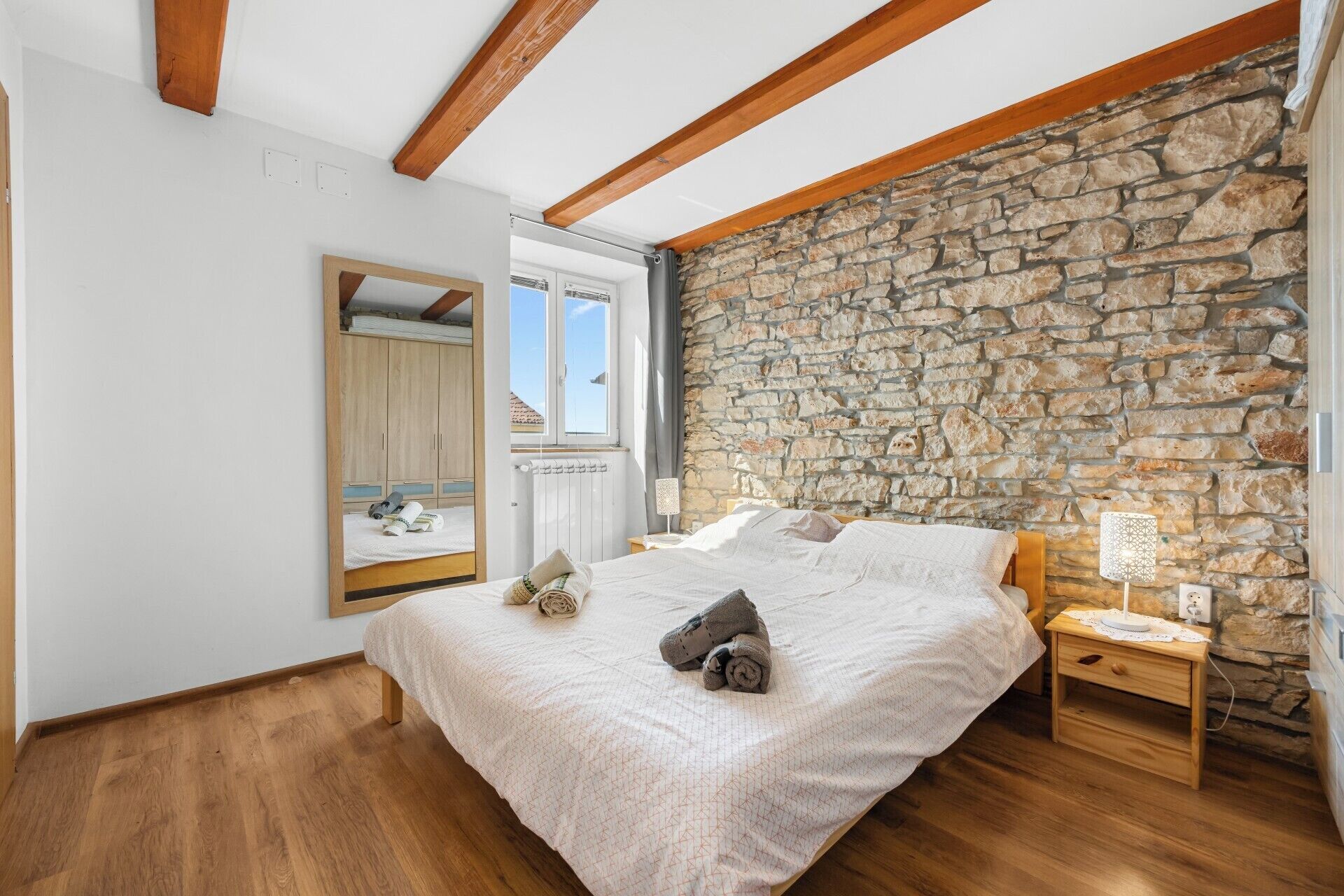 A bedroom with a stone wall, wooden beams, a bed with towels, a nightstand, and a window.