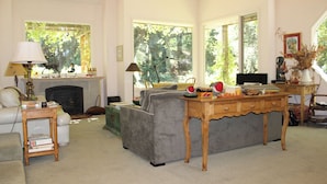 A living room with a fireplace, a sofa, a wooden table, and large windows with a view of trees.