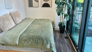 Quarto com cama, almofadas, mesa de cabeceira, luminária, obra de arte na parede e planta em vaso.