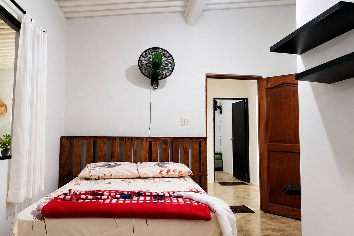 Quarto com cama, ventoinha na parede, cabeceira de madeira, porta e espelho.
