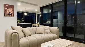 A modern living room with a sofa and a coffee table, a kitchen area in the background, and a large sliding glass door. - The Webster in the City (Reid)
