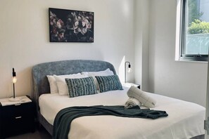 Ein ordentlich eingerichtetes Schlafzimmer mit Bett, Kissen, einer Nachttischlampe, einem Wandbild und einem Fenster.