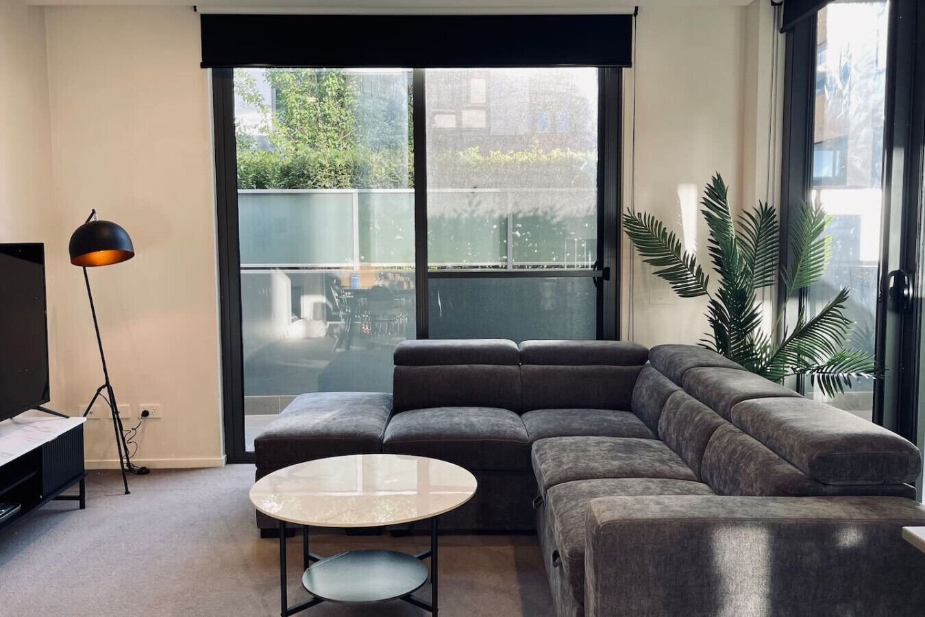 Een moderne woonkamer met een hoekbank, een ronde tafel, een vloerlamp en een groot raam met uitzicht op groen.