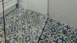 A bathroom with a shower, a glass door, a white tiled wall, and patterned floor tiles.