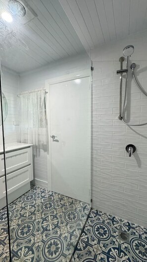 Un baño con ducha, puerta de vidrio, pared de azulejos blancos y pisos con diseño.
