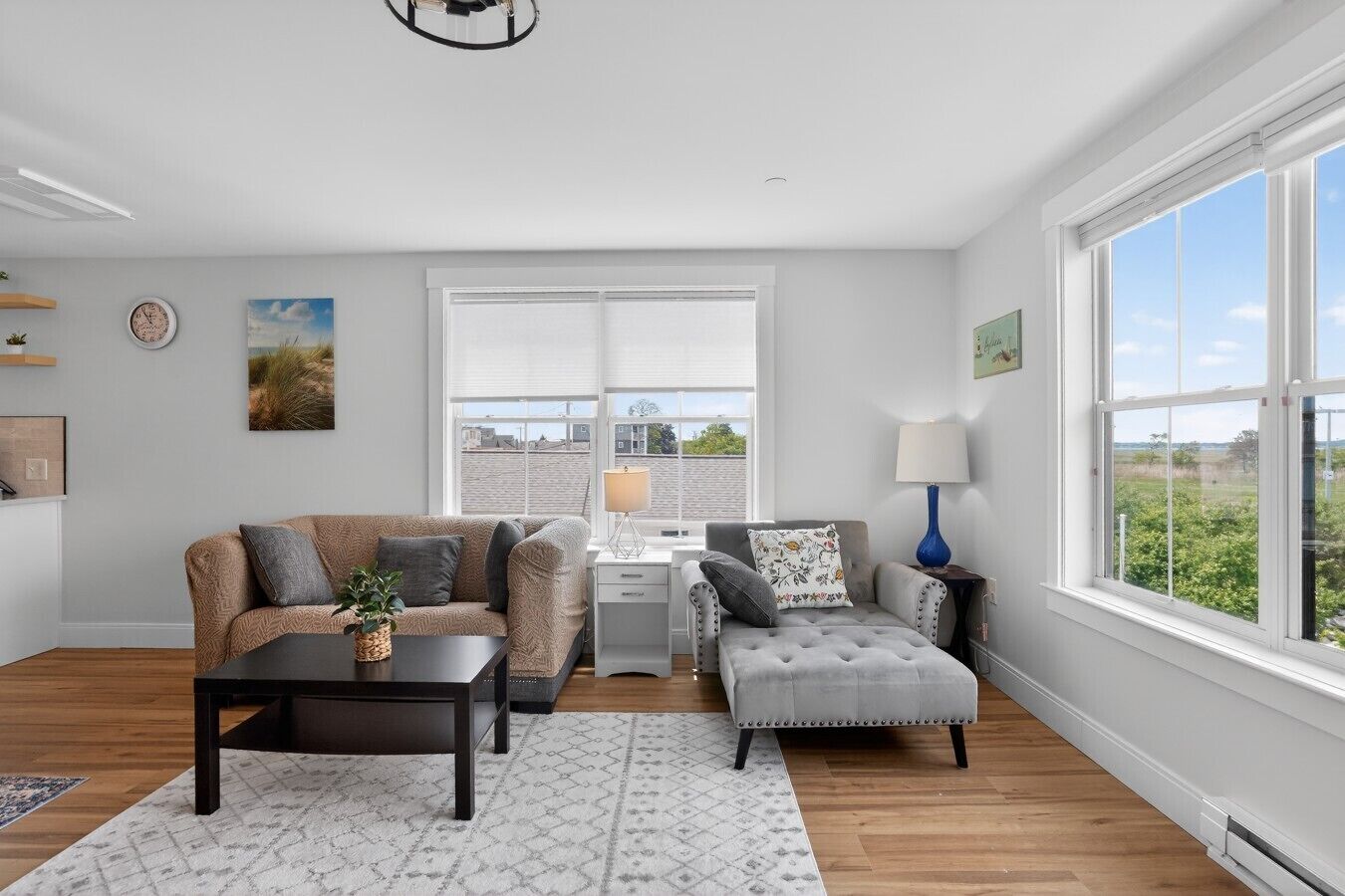 Een moderne woonkamer met een bank, een fauteuil, een salontafel, een bureau en een lamp. Er is een groot raam met uitzicht op buiten.