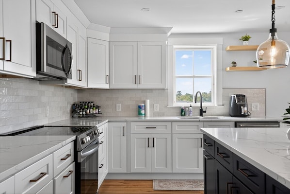 Una cocina moderna con armarios blancos, estufa de acero inoxidable, microondas, cafetera y ventana que ofrece luz natural.