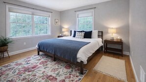 A bedroom with a bed, two bedside tables, a nightstand, a potted plant, a floral rug, and two windows with blinds. - Sleep up to 12, quick access to Kittery and Portsmouth, Beaches, Boat Launch (Eliot)