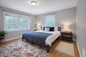 Un dormitorio con una cama, dos mesitas de noche, una lámpara auxiliar, una planta en maceta, una alfombra estampada y dos ventanas con persianas.
