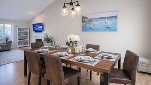 A dining area with a wooden table set for four, surrounded by chairs, a painting of a coastal scene, and a television on the wall. - Sleep up to 12, quick access to Kittery and Portsmouth, Beaches, Boat Launch (Eliot)