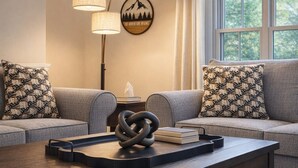 A living room with two grey sofas, patterned throw pillows, a wall-mounted lamp, a framed picture, and a coffee table with a coiled metal sculpture and books. - Sleep up to 12, quick access to Kittery and Portsmouth, Beaches, Boat Launch (Eliot)