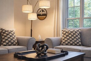 A living room with two grey sofas, patterned throw pillows, a wall-mounted lamp, a framed picture, and a coffee table with a coiled metal sculpture and books. - Sleep up to 12, quick access to Kittery and Portsmouth, Beaches, Boat Launch (Eliot)