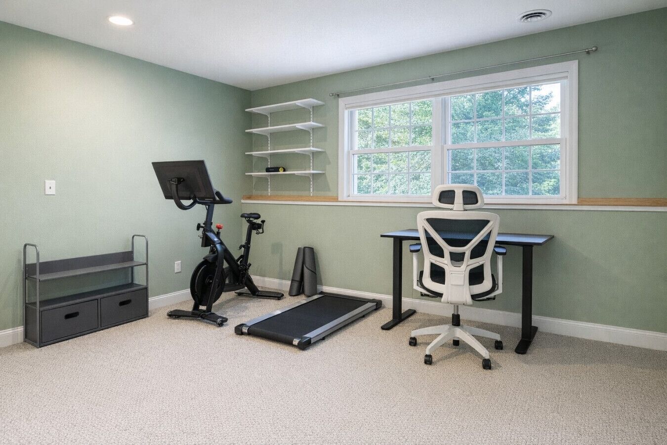 Ein Zimmer mit Laufband, Ergometer, Schreibtisch mit Stuhl und Fenster mit Blick auf Bäume.