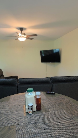 Una sala de estar con ventilador de techo, un televisor de pantalla plana montado en la pared, un sofá oscuro y una mesa de centro con una jarrón de leche, un frasco de pimienta negra y un control remoto.