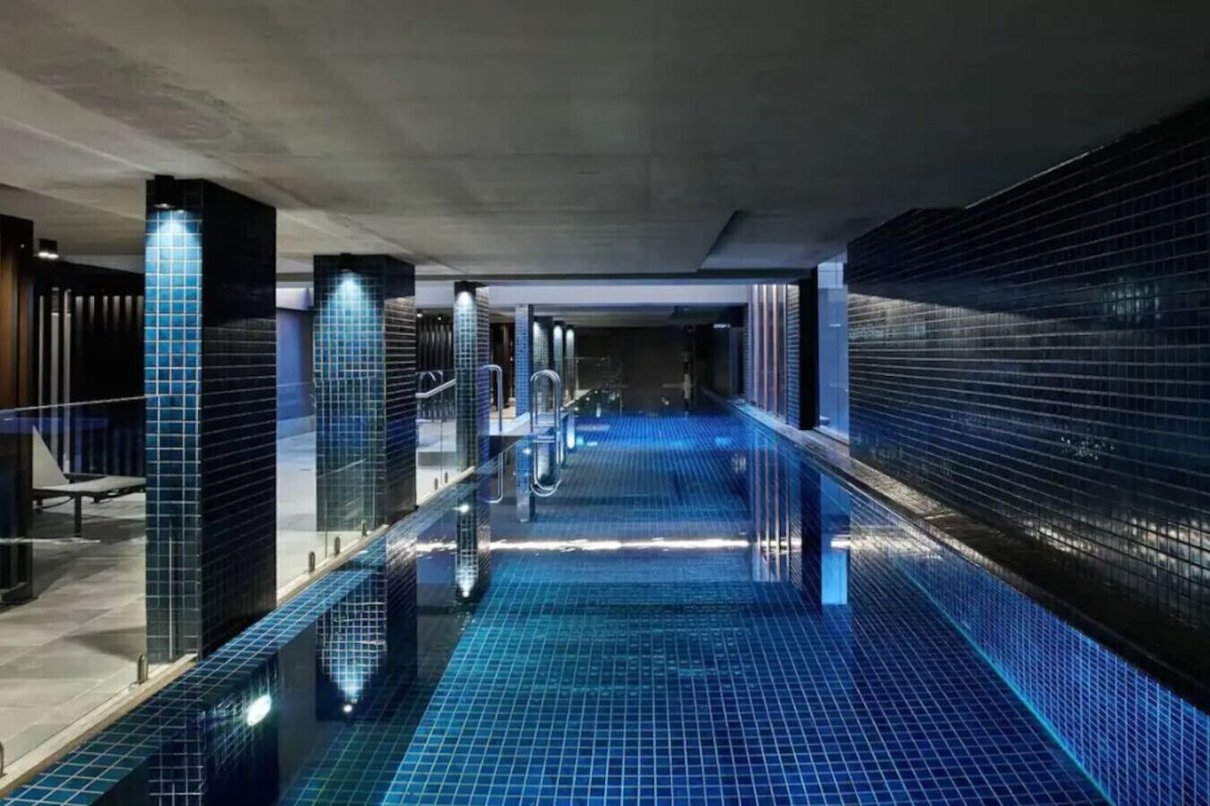 A modern indoor pool with a glass barrier, tiled floor, and a concrete ceiling.
