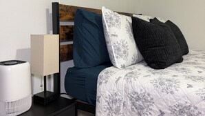 A neatly arranged bedroom with a bed, a bedside table with a lamp, and a wall-mounted shelf.