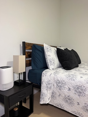 A neatly arranged bedroom with a bed, a bedside table with a lamp, and a wall-mounted shelf.