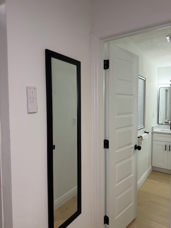 A hallway with a door, a mirror, and a bathroom with a sink and cabinets.