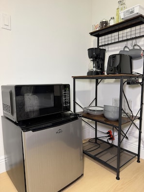 A compact kitchen with a microwave, a stainless steel mini-fridge, a coffee maker, and a shelf with kitchenware.