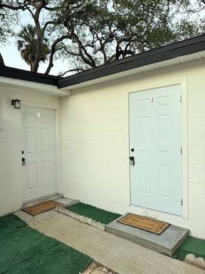 A building with two numbered doors, a green mat at the entrance, and a tree in the background.