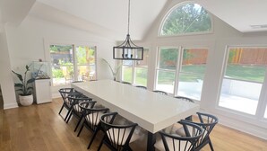 Un comedor amplio con una larga mesa blanca y sillas negras, ventanas grandes y una lámpara colgante.