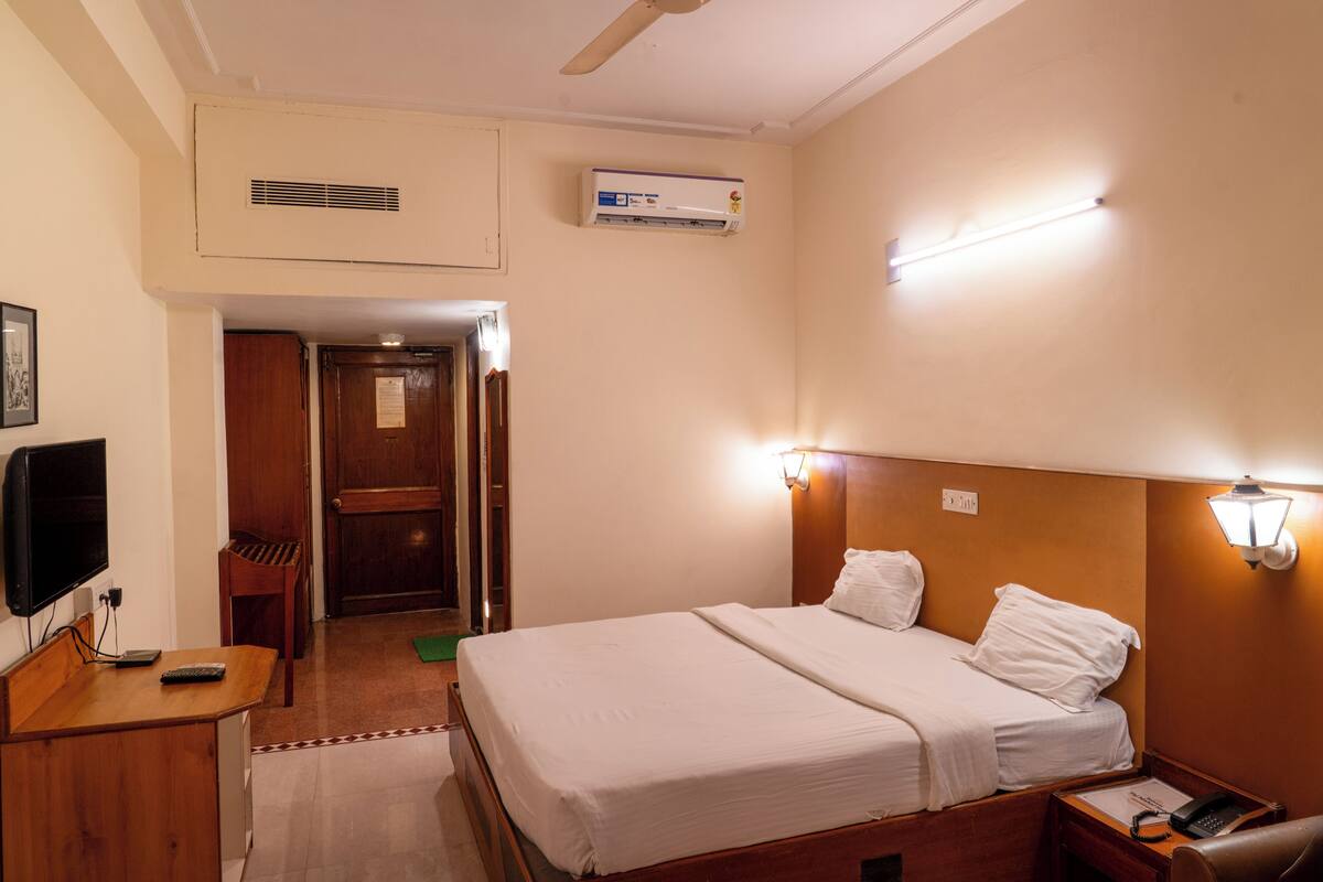 Habitación de hotel con cama, mesita de noche, lámpara, aire acondicionado y televisión.
