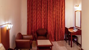 A hotel room with a large window, a ceiling fan, a wooden table, a chair, a lamp, and a mirror. - Belsons TajMahal Hotel (Hyderabad)