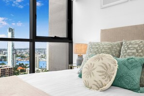 Ein Hotelzimmer mit großem Fenster, einem Bett mit gemusterten Kissen, einer Nachttischlampe und Blick auf eine Stadtlandschaft.