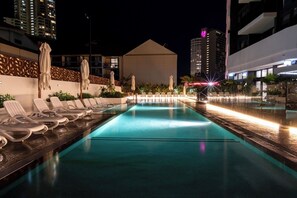 Ein Pool bei Nacht mit Liegestühlen und Sonnenschirmen, umgeben von Gebäuden.