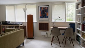 A room with a sofa, a desk with two chairs, a bookshelf, a radiator, and a window with blinds.