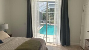 Ein Schlafzimmer mit Bett, Nachttisch, Lampe, einem grünen Zierkissen und Blick auf einen Swimmingpool durch eine Glastür.
