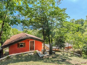 Una cabaña con techo rojo, puerta de madera y contraventanas, rodeada de abundante vegetación y árboles.