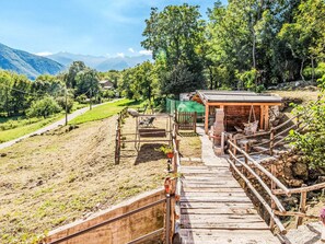 Un escalier en bois mène à une structure rustique également en bois, agrémentée d’une rambarde en métal, et l’ensemble est entouré d’une zone herbeuse et d’arbres.