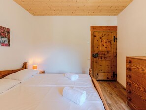 Quarto com duas camas, cômoda de madeira, porta de madeira e luminária fixa na parede.