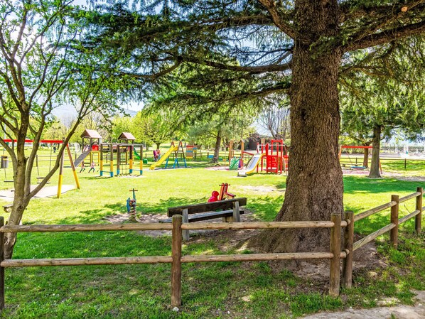 Un parque infantil con diversas estructuras de juego, un banco y un árbol grande al frente.
