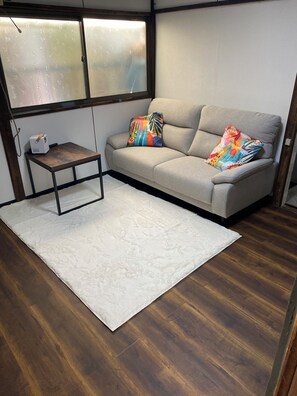 Ein Zimmer mit einer Couch, einem kleinen Tisch, einem weißen Teppich, Holzböden und einem Fenster mit gesinterten Scheiben.