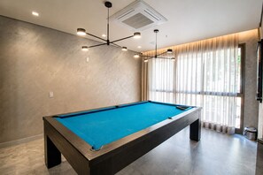 Ein Billardtisch in einem Raum mit einer modernen Deckenleuchte und großen Fenstern mit Vorhängen.