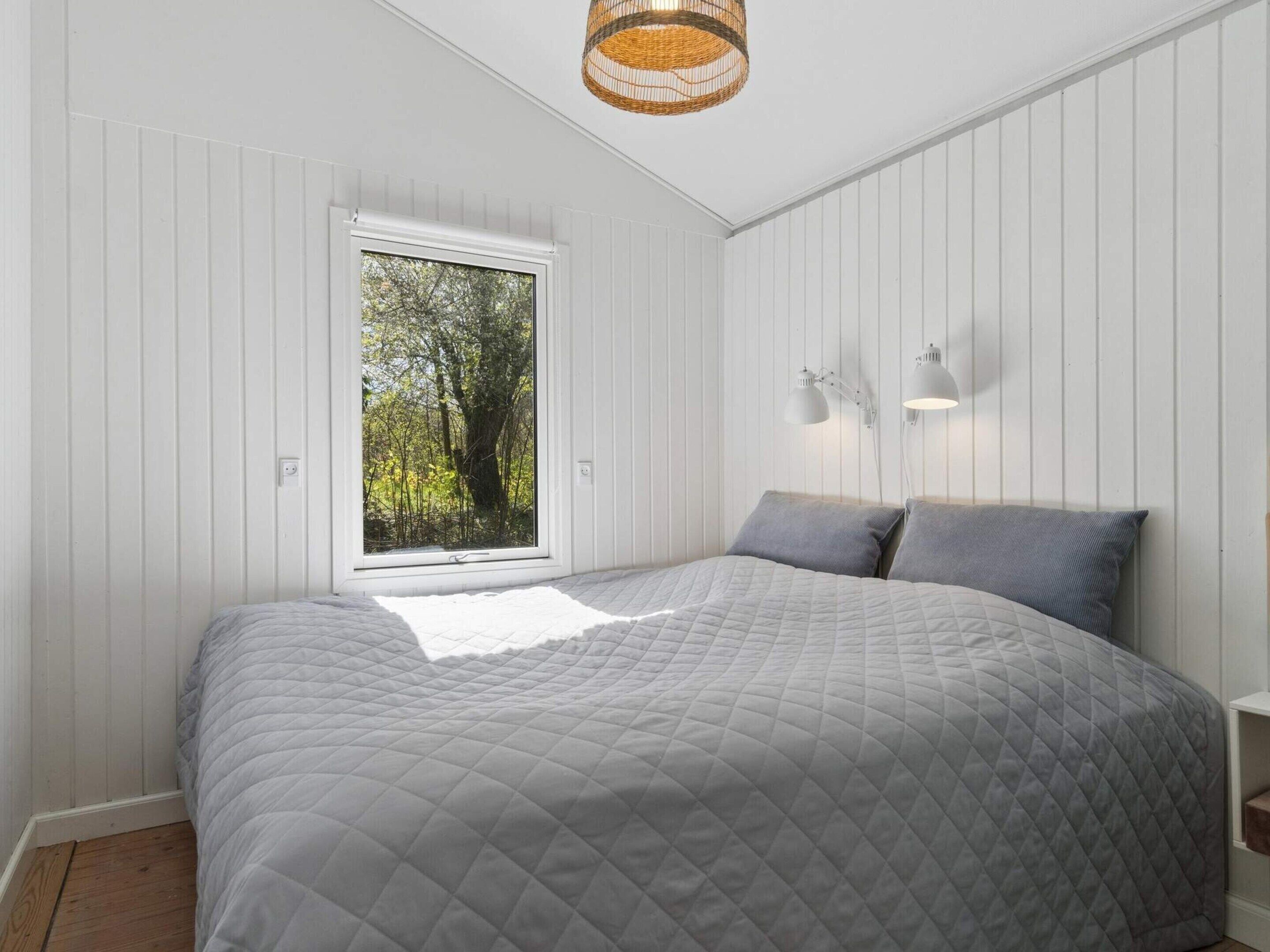 Ein Schlafzimmer mit einem großen Bett, einem Fenster mit Blick auf Bäume, weißen Wandverkleidungen und einer hängenden Leuchte.