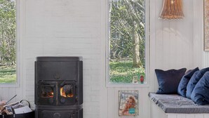 A room with a wood-burning stove, a window with a view of trees, a bench with cushions, and a wall-mounted light fixture. - 4 Star Holiday Home in Spottrup (Spøttrup)