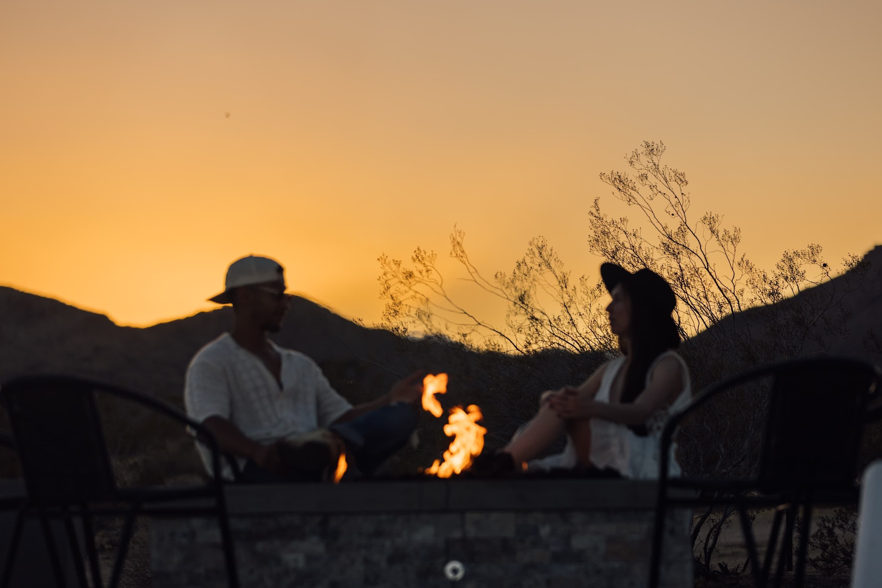 Two people sitting on a bench with a fire between them, in a setting with a stone wall and sparse vegetation.