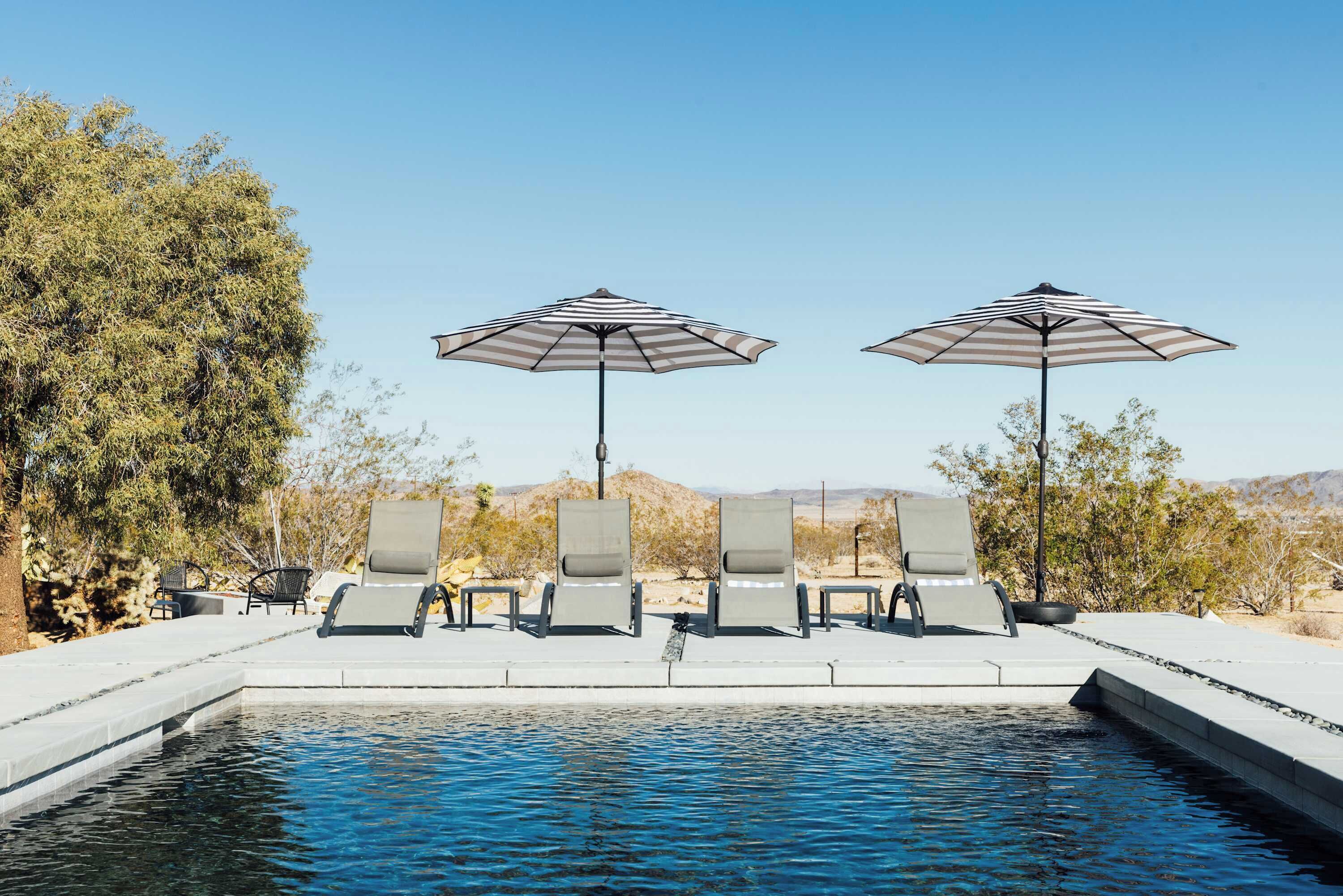Zona de piscina con sillones de descanso y sombrillas a rayas, rodeada de árboles y un cielo despejado.