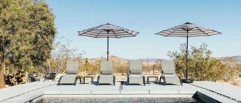 Zona de piscina con sillones de descanso y sombrillas a rayas, rodeada de árboles y un cielo despejado.