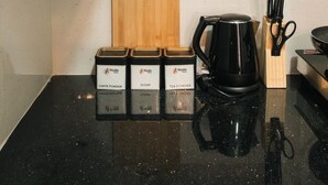 Una cocina con una cafetera negra, una tabla de cortar de madera y tres recipientes de especias etiquetados como café en polvo, azúcar y té en polvo.