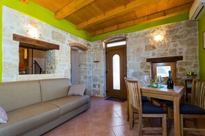 Ein gemütliches Zimmer mit Steinwand, Holzboden, einer Couch, einem Esstisch mit Stühlen und einer Tür zu einem anderen Raum.